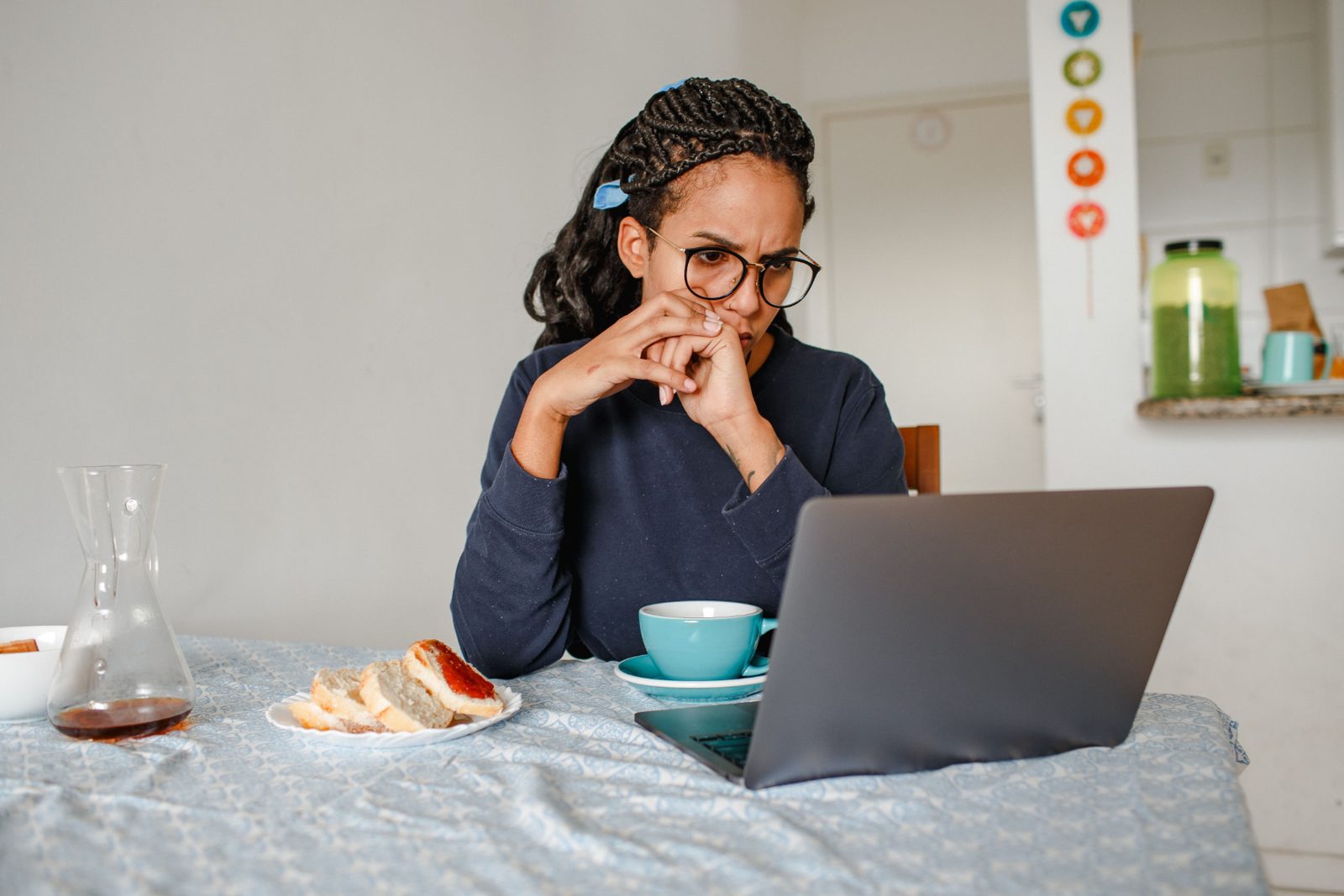 Woman with glasses and braided hair sitting at her kitchen table, hands to her mouth, looking at her laptop next to coffee and toast