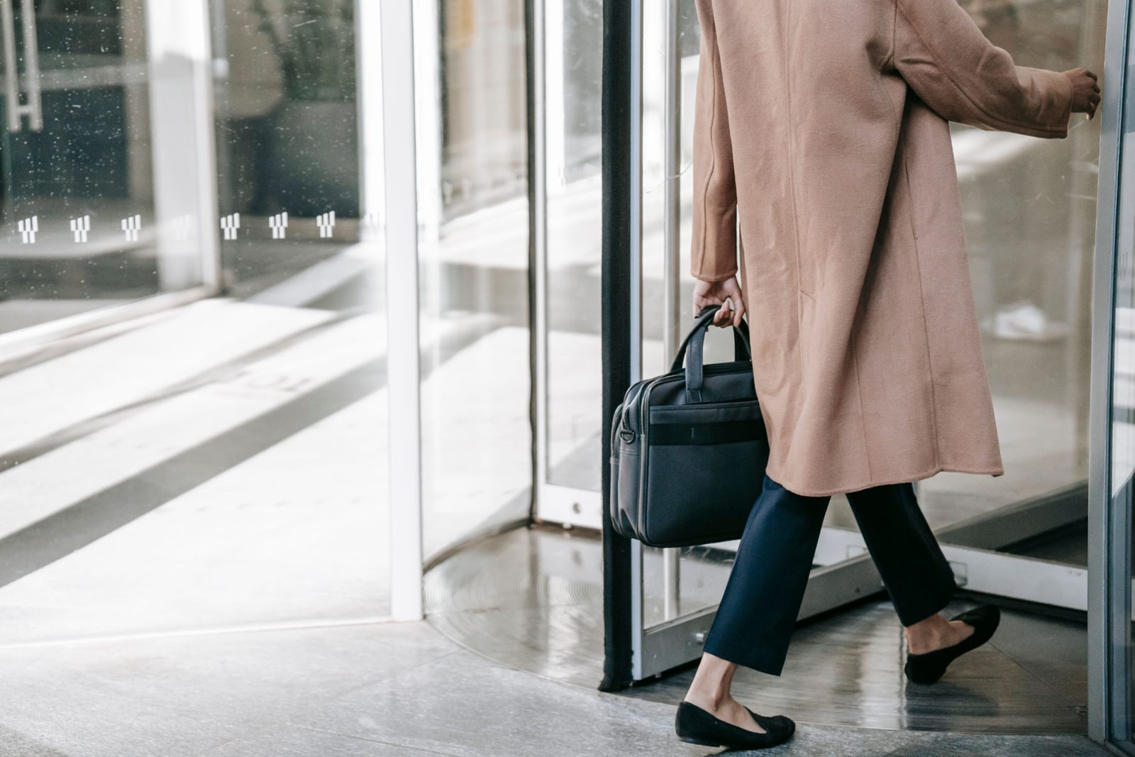 Woman in a camel coat carrying a laptop bag stepping through the revolving door of an office building