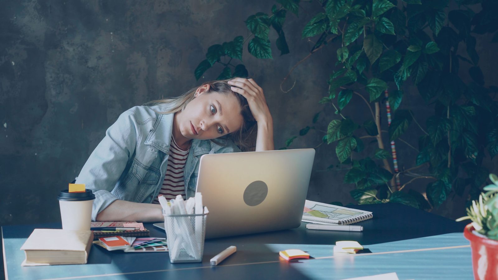 Woman resting her head on her hand at a desk, looking tired at her laptop amid notebooks, markers and a coffee cup