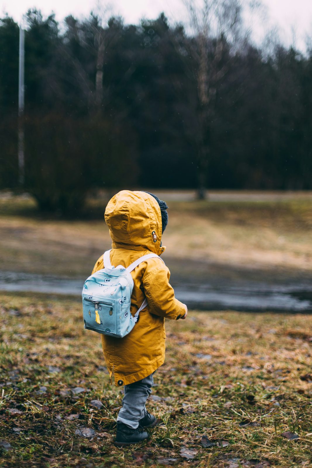 Young child in a yellow raincoat with a light blue backpack walking outdoors across a grassy field
